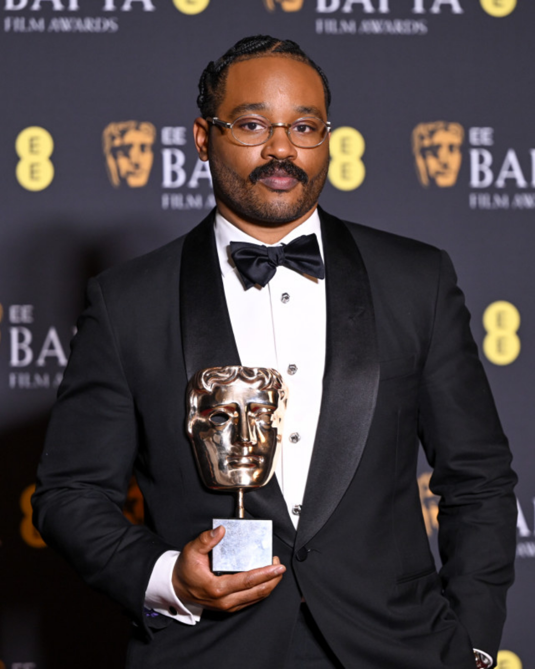 Ryan Coogler posando con un esmoquin negro y el premio BAFTA
