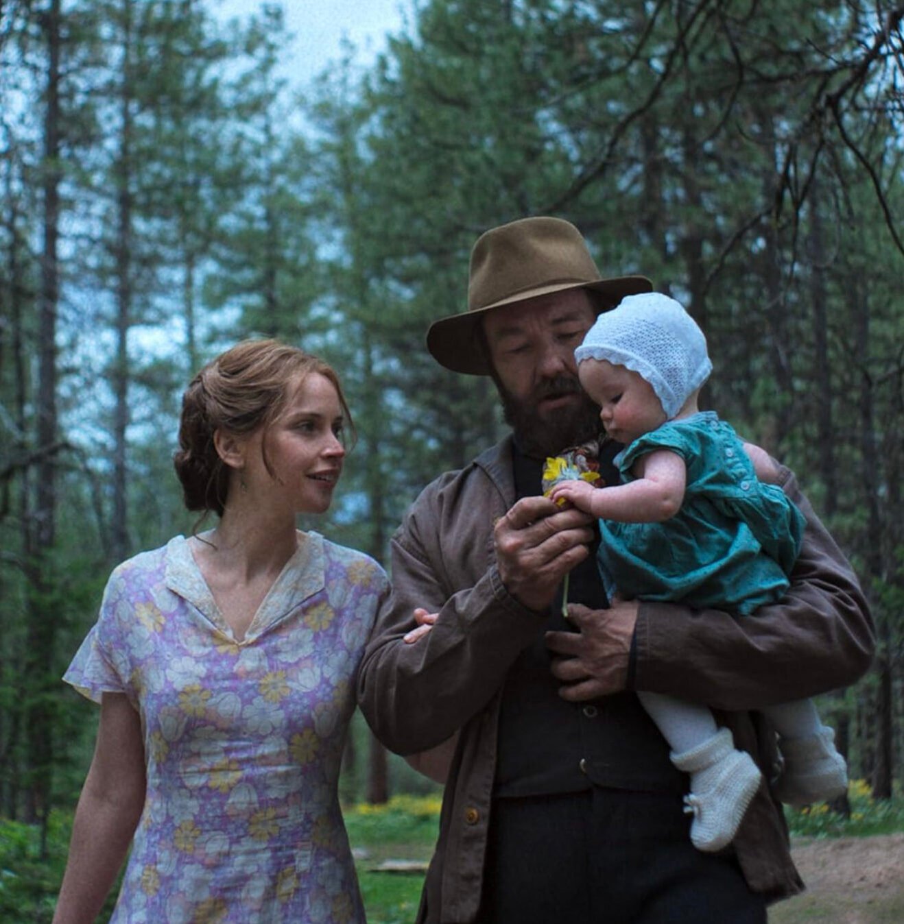 Joel Edgerton con sombrero carga a un bebé mientras Felicity Jones camina a su lado sonriendo por un bosque en una escena de 'Train Dreams'.