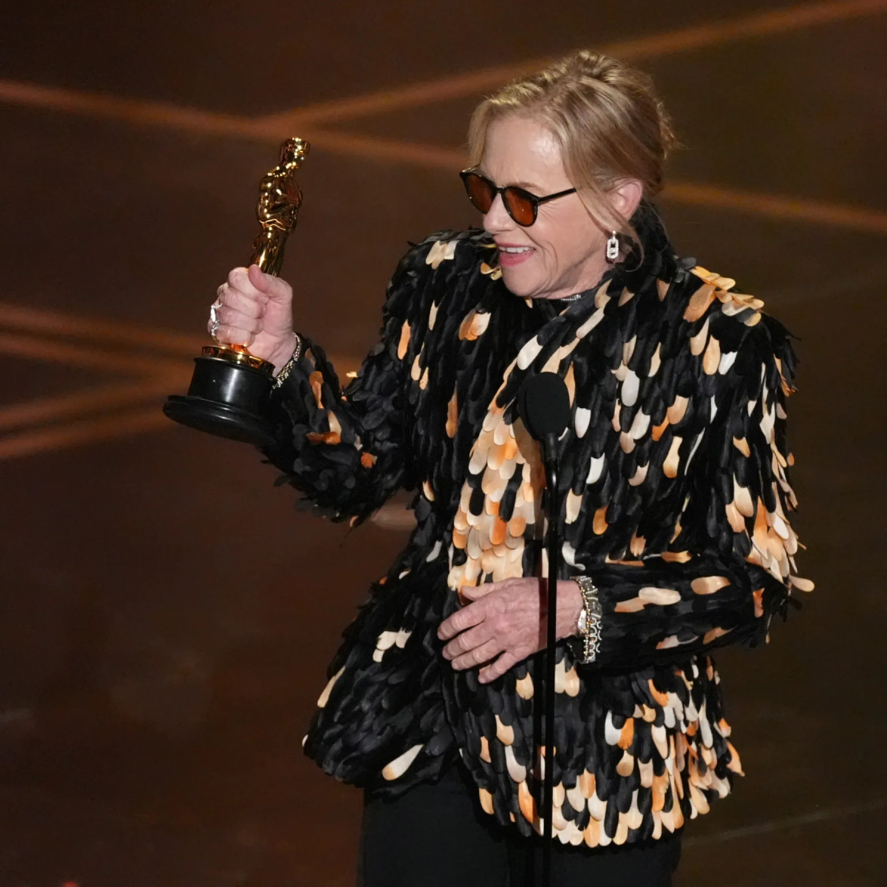 Amy Madigan en el escenario de los Óscar 2026, sostiene la estatuilla dorada del Óscar en su mano derecha, levantándose ligeramente con una expresión de alegría y sorpresa

